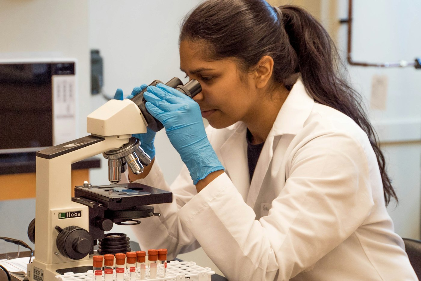 Scientist in a white lab coat and blue gloves looking through a microscope